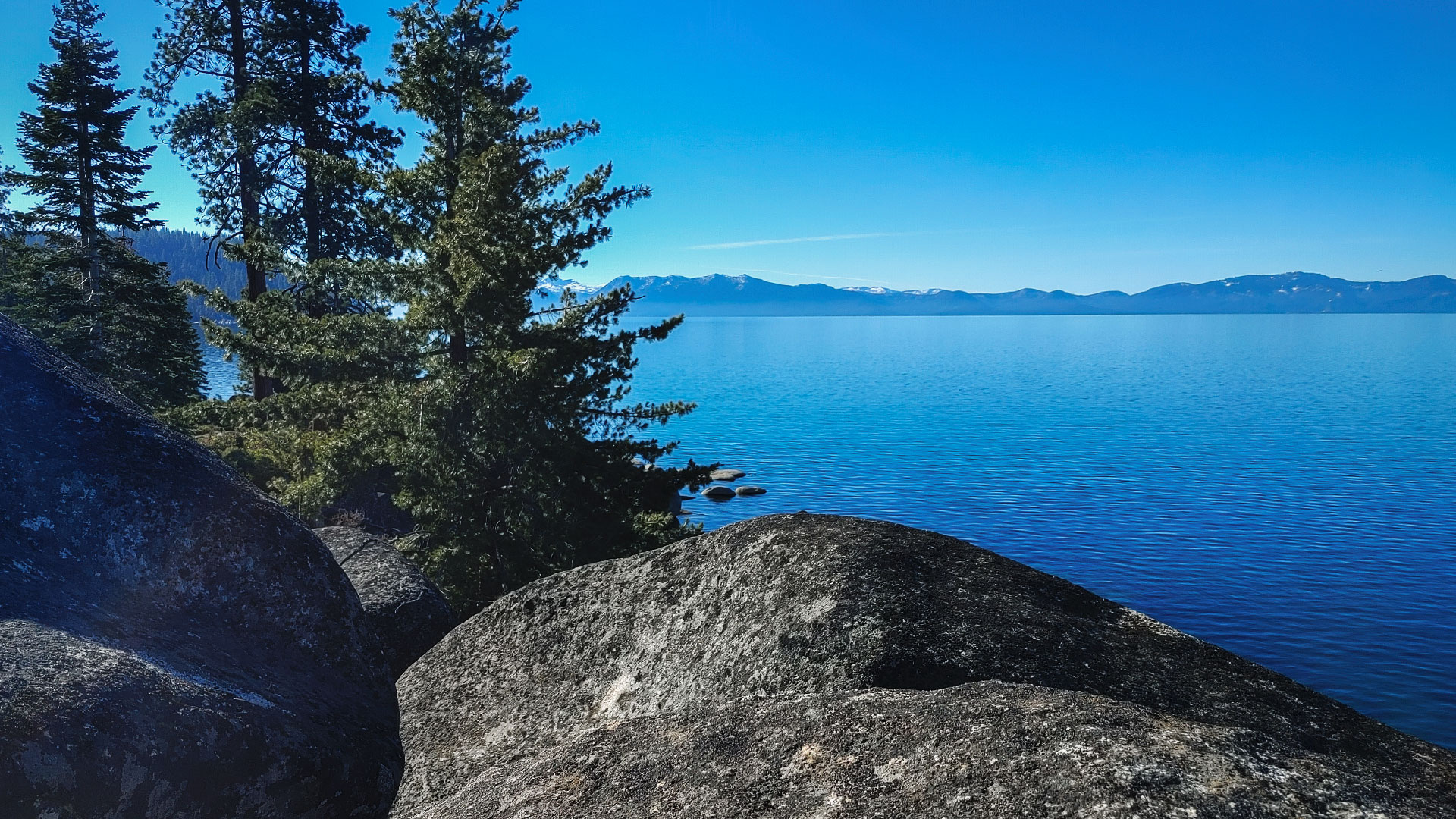 Blue skies and blue water with granite boulders and pine trees in the foreground